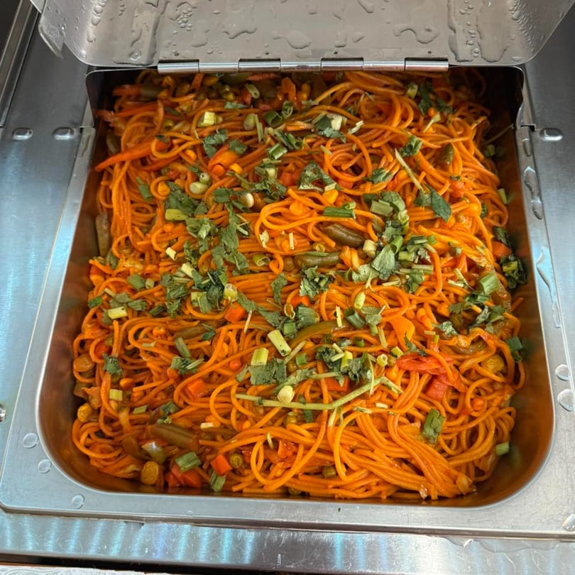 A large metal tray filled with orange noodles, vegetables, and green herbs, ready to be served.